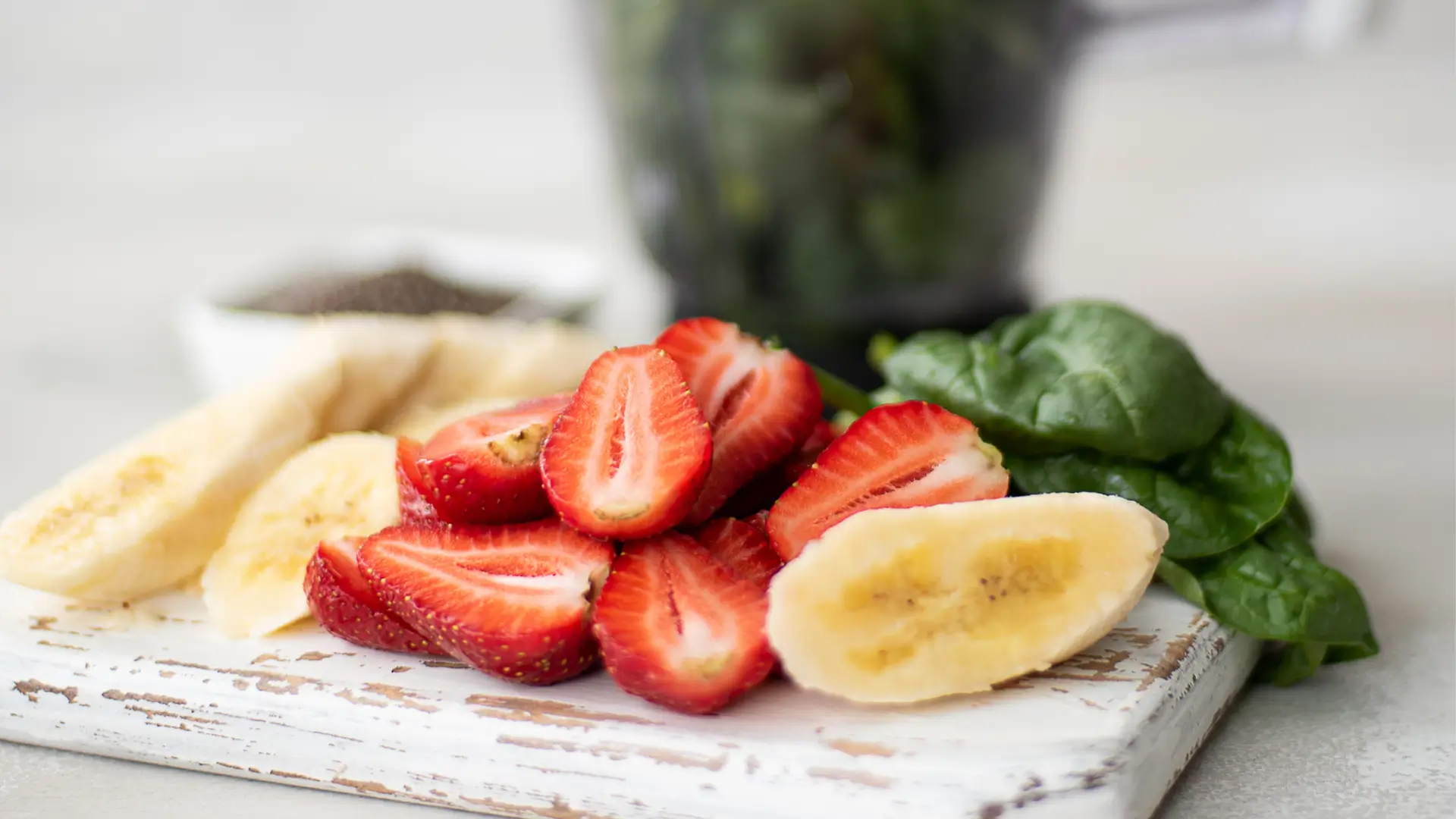 Smoothie ingredients on a counter: a carton of oat milk, a bowl of spinach, a bowl of frozen berries, and sliced bananas on a cutting board, with a window view in the background.