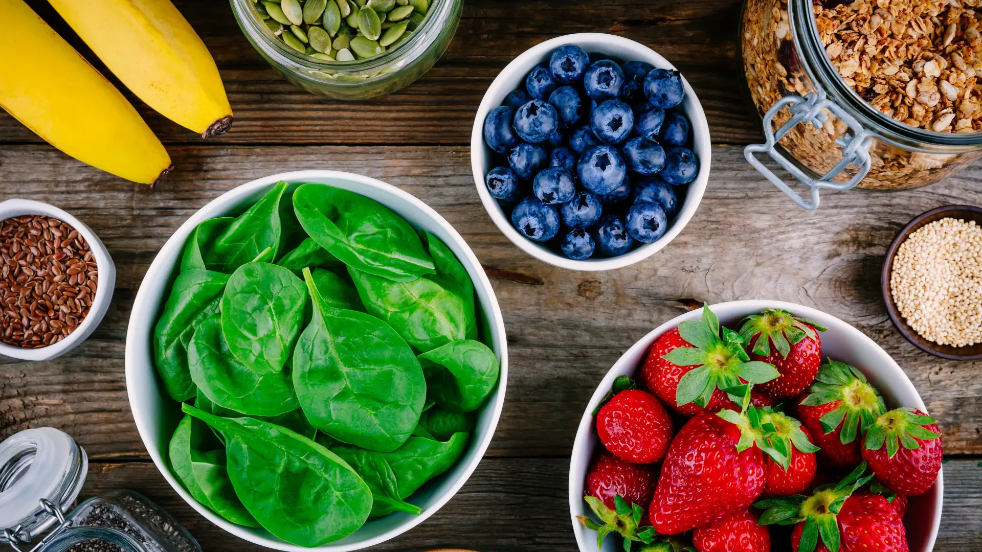 Smoothie ingredients on a counter: a carton of oat milk, a bowl of spinach, a bowl of frozen berries, and sliced bananas on a cutting board, with a window view in the background.