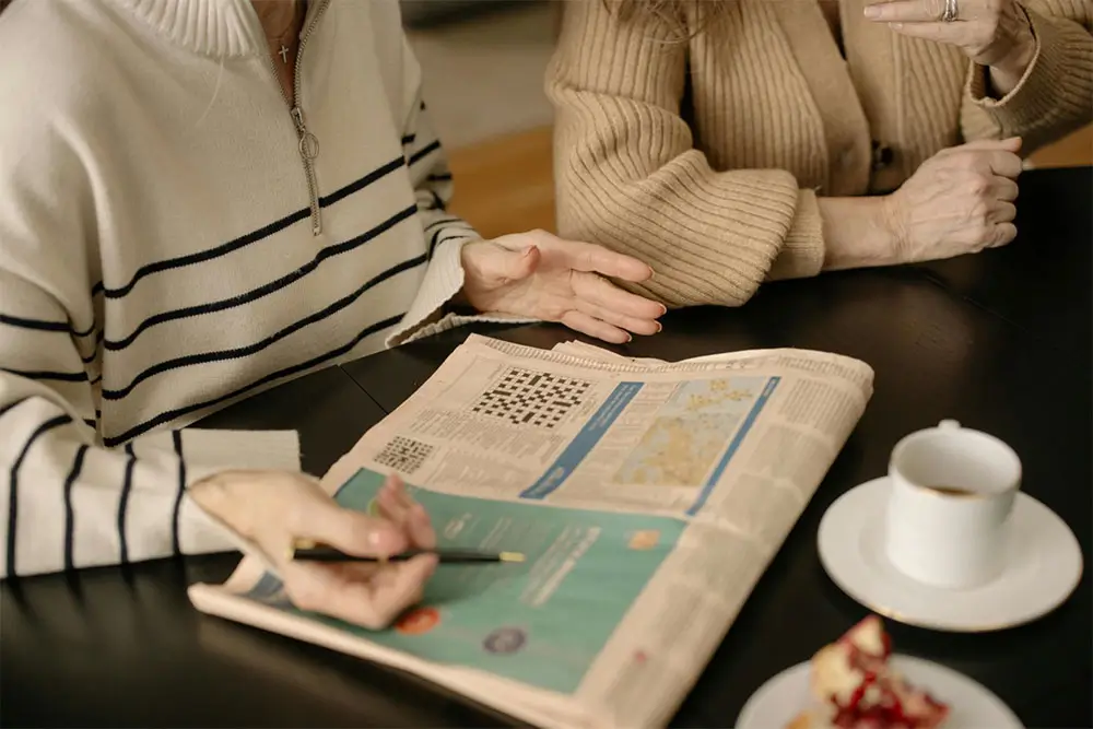 Two individuals sitting at a table engaged in solving a crossword puzzle from a newspaper, with a cup of coffee and a slice of cake nearby, creating a cozy and interactive atmosphere.